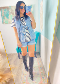 Woman taking a mirror selfie wearing a striped shirt, denim vest, shorts, and knee-high boots in a room with a colorful rug.