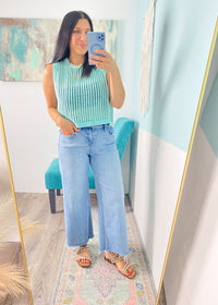 Woman taking a mirror selfie wearing  mint green sleeveless crochet top  and jeans in a room with a colorful rug and wall art.