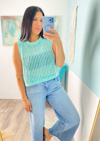 Woman taking a mirror selfie wearing a mint green sleeveless crochet top and jeans in a room with light blue walls.