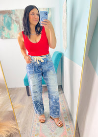 Woman taking a mirror selfie wearing a red lace patterned seamless cropped top and blue jeans in a room with colorful decor.