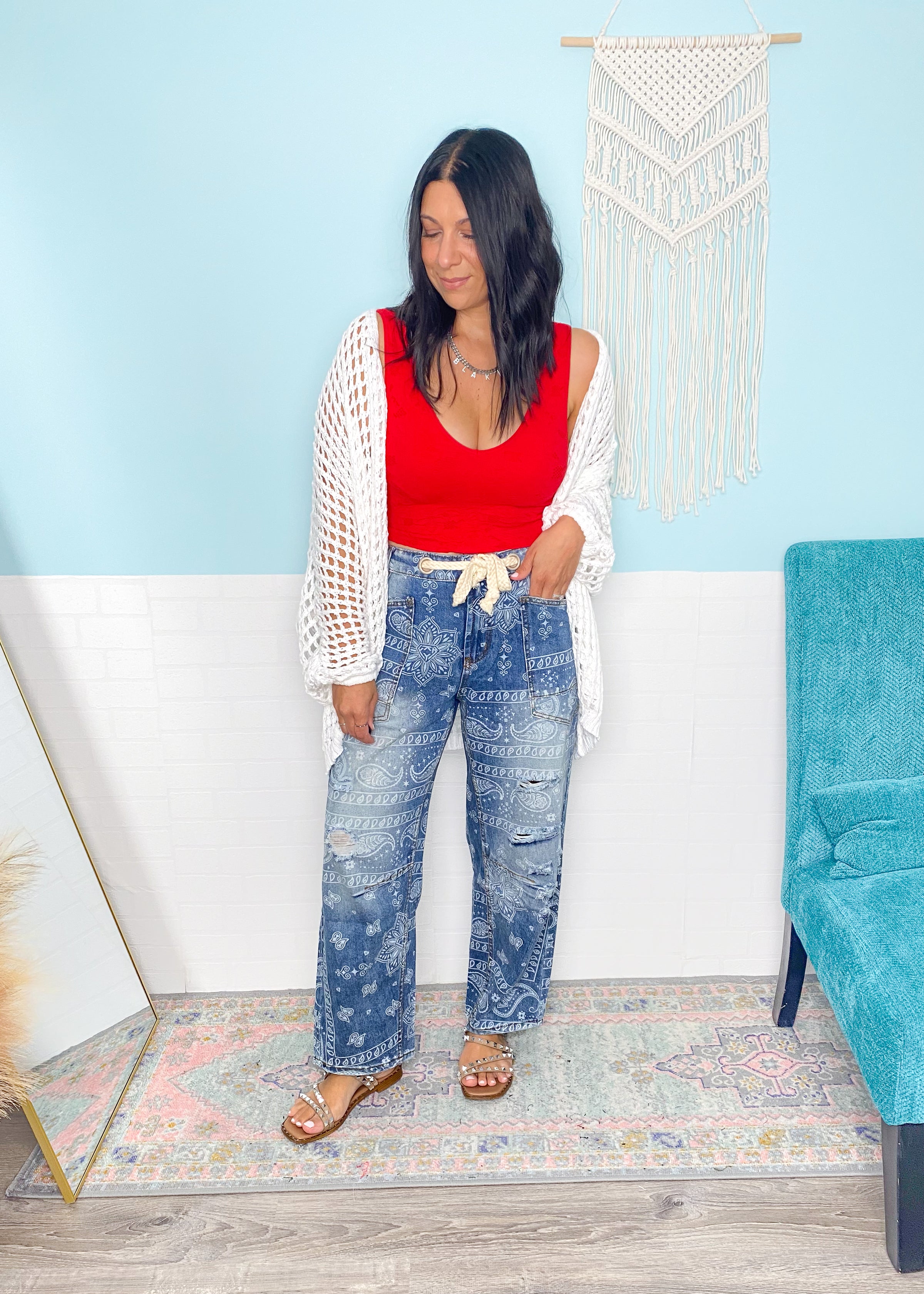 Woman in a red top with a crochet ivory cardigan and blue jeans standing in a room with a light blue wall and teal chair.