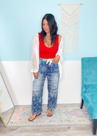 Woman wearing a red top and paisley printed jeans blue jeans standing in a room with a blue wall and a turquoise chair.