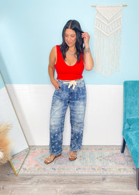 Woman in a red lace patterned seamless cropped top and blue jeans standing in a room with a white wall and decorative elements.