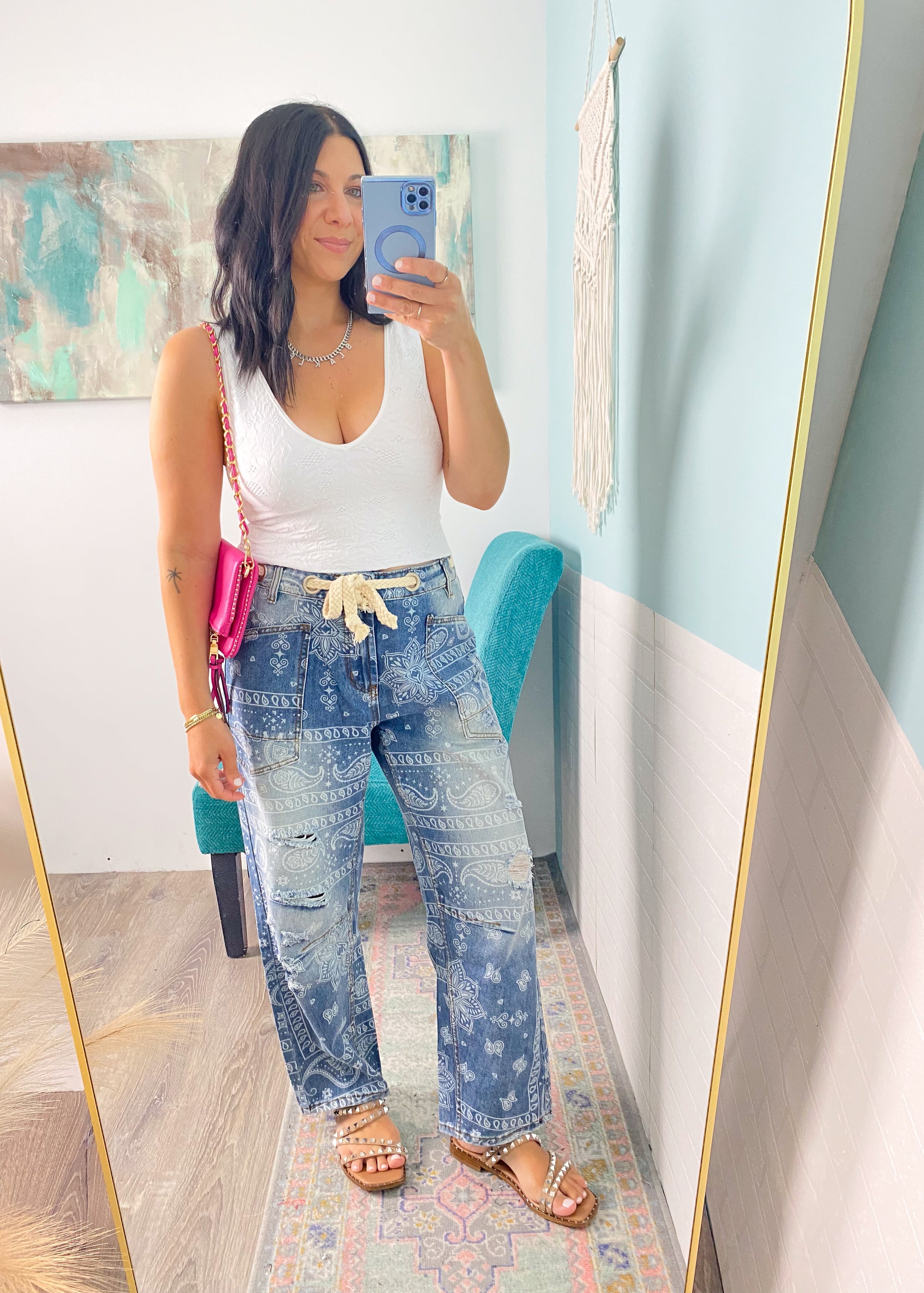 Woman taking a mirror selfie wearing a white tank top and paisley printed jeans in a room with colorful decor.