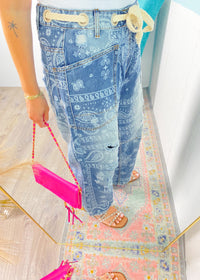 Person wearing blue bandana print pants with a pink bag on a colorful rug.