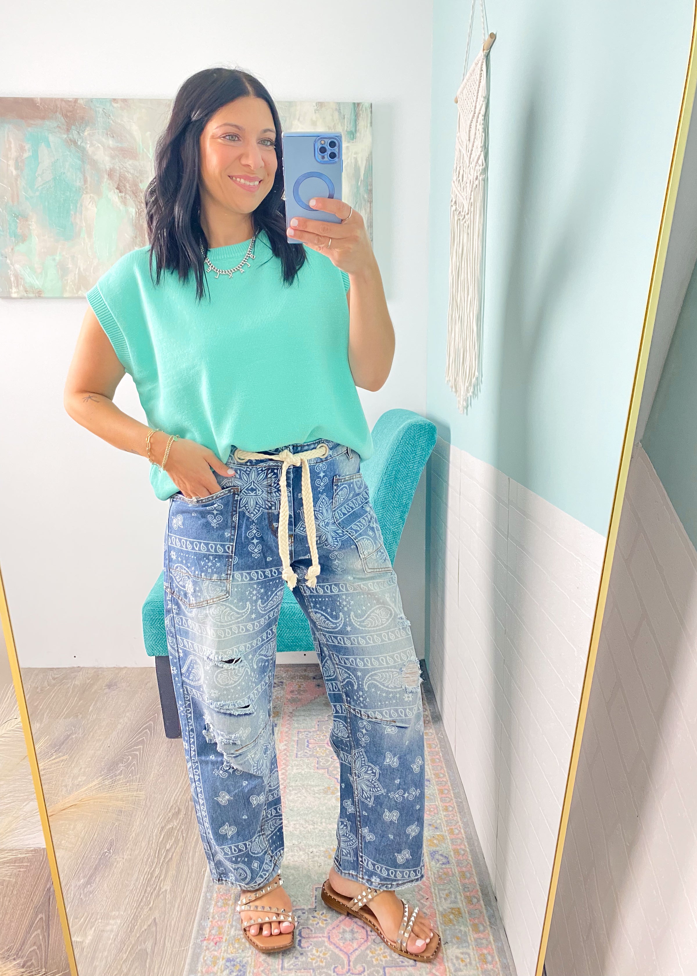 Woman taking a mirror selfie wearing a mint green top and paisley printed blue jeans in a room with light blue walls.