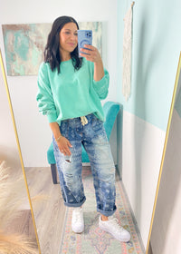 Woman taking a mirror selfie wearing a green sweatshirt and paisley printed blue jeans indoors.