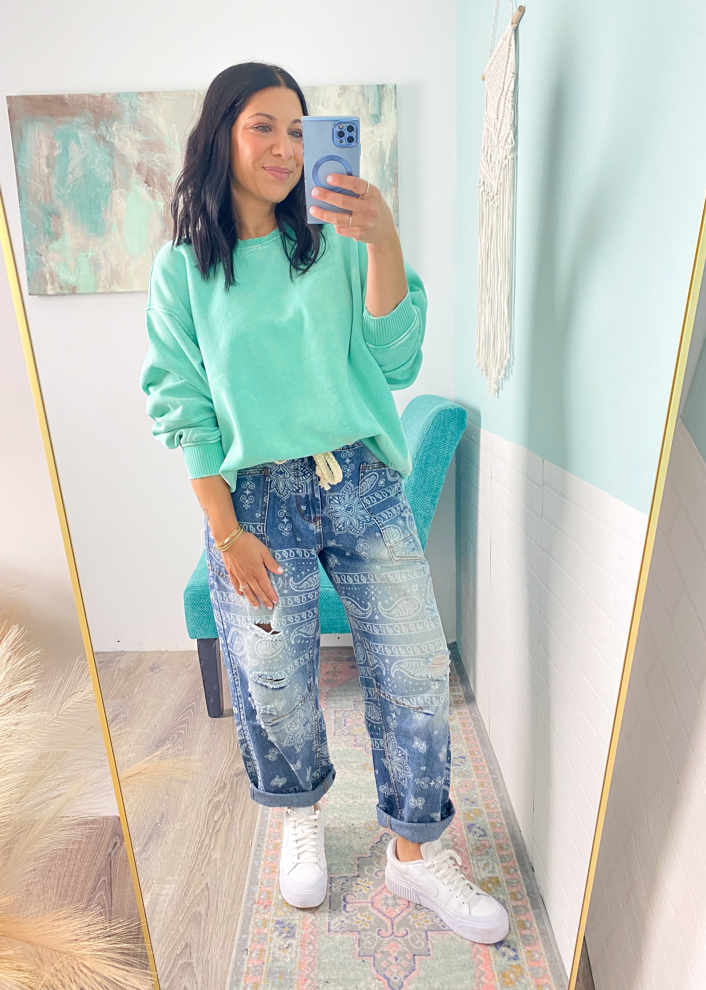 Woman taking a mirror selfie wearing a green sweatshirt and paisley printed blue jeans indoors.