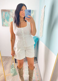 Woman taking a mirror selfie wearing an off white peplum top and off white shorts outfit with beige boots in a room with light blue walls.