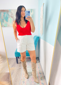 Woman taking a mirror selfie wearing a red lace patterned seamless cropped top, white shorts, and beige knee-high boots in a room with a colorful rug and abstract painting.