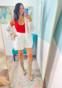 Person taking a mirror selfie wearing a white lace cardigan, red top, white shorts, and beige knee-high boots.