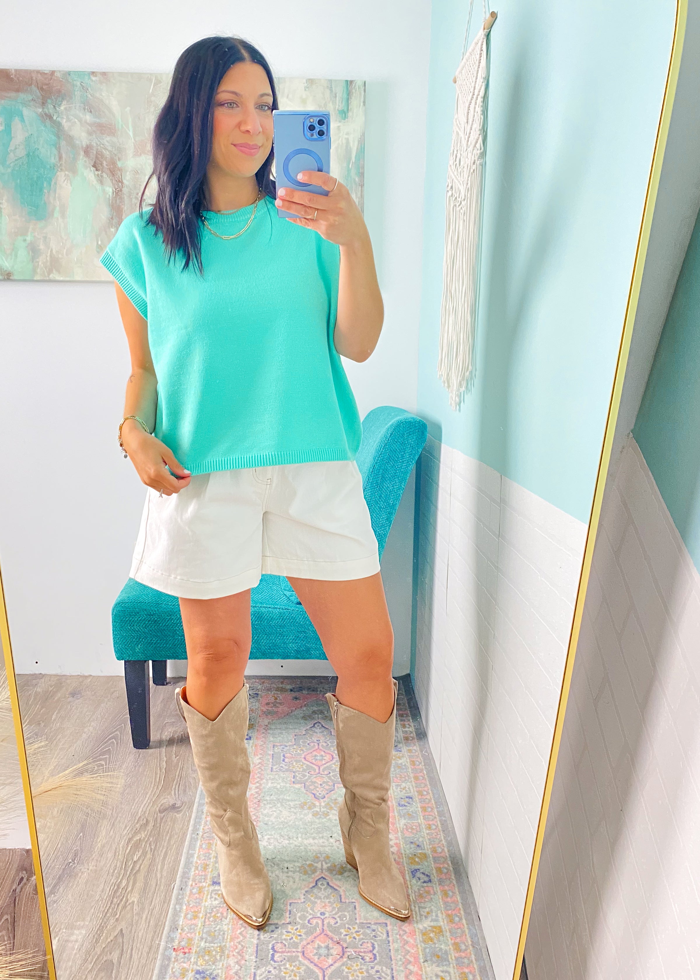 Woman taking a mirror selfie wearing a mint green top, white shorts, and beige knee-high boots in a room with a colorful rug and abstract painting.