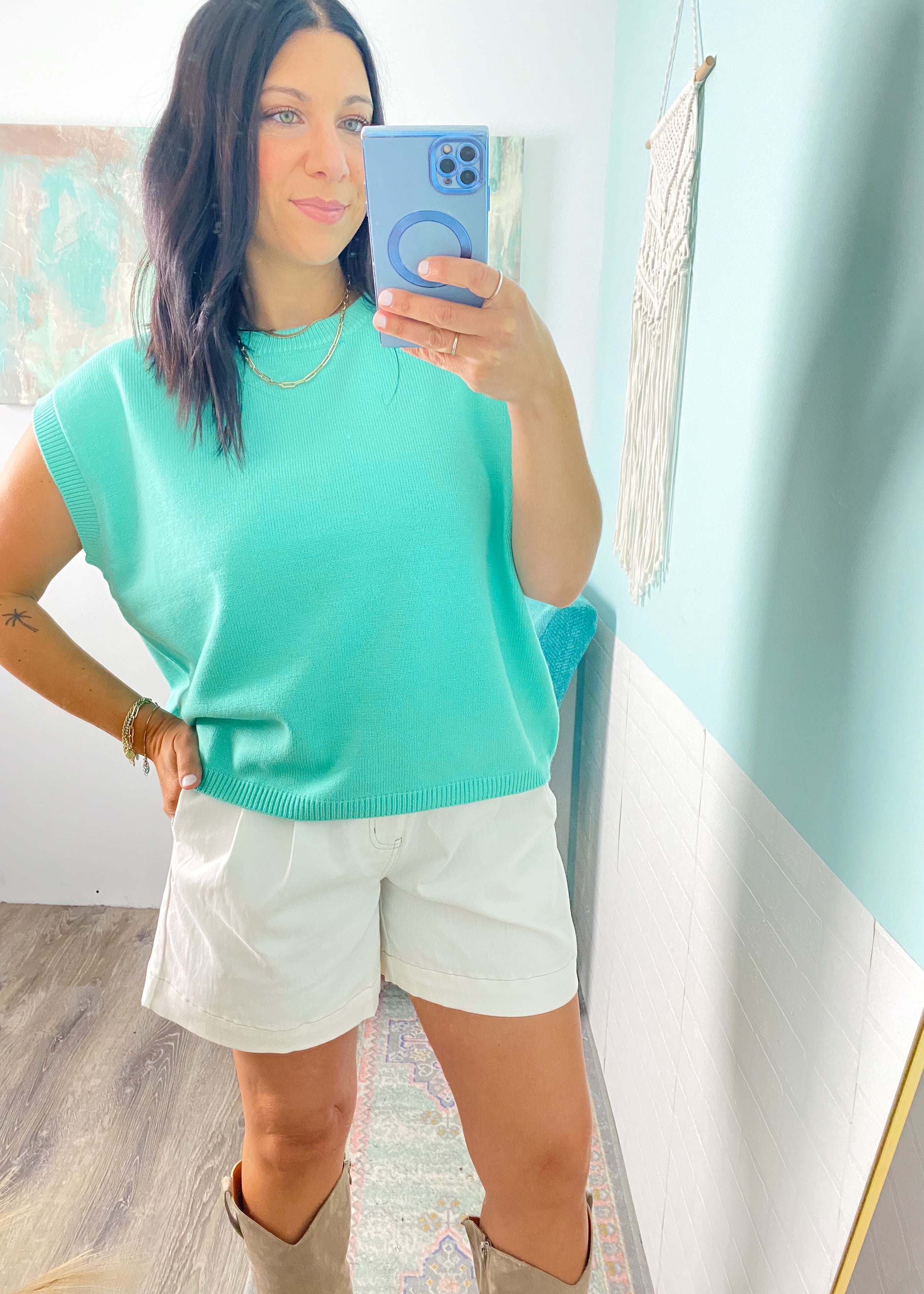 Woman taking a mirror selfie wearing a mint green top and white shorts in a room with light blue walls.