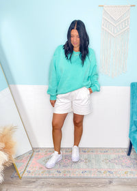 Woman wearing a mint green sweatshirt and white shorts standing in a room with a light blue wall and decorative rug.