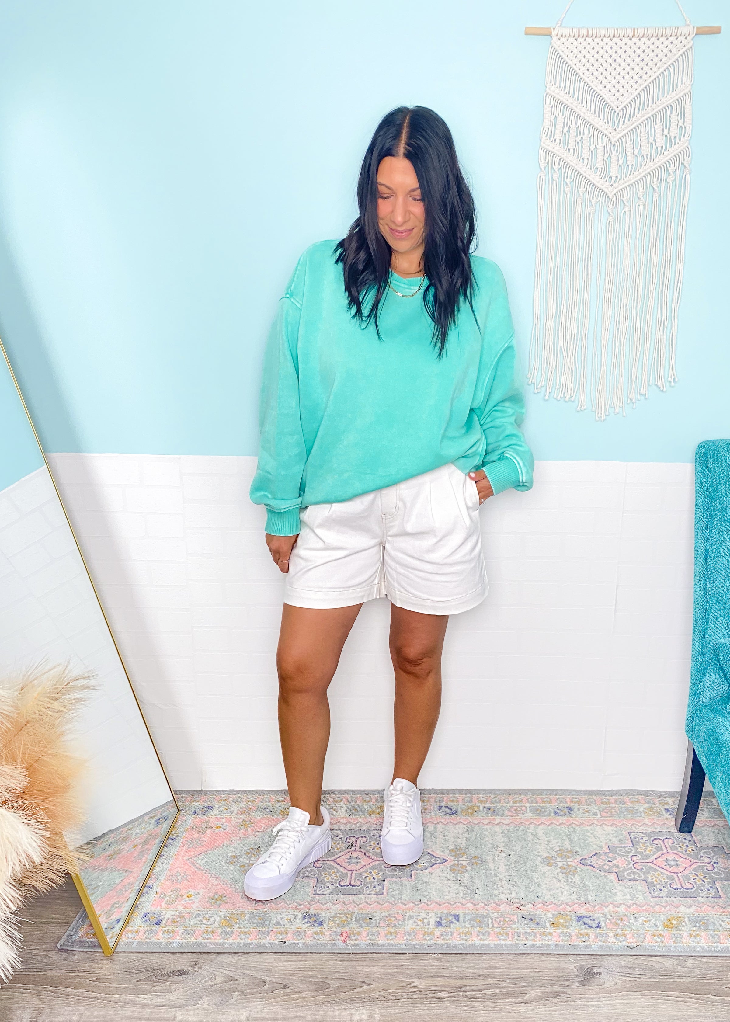 Woman wearing a mint green sweatshirt and white shorts standing in a room with a light blue wall and decorative rug.