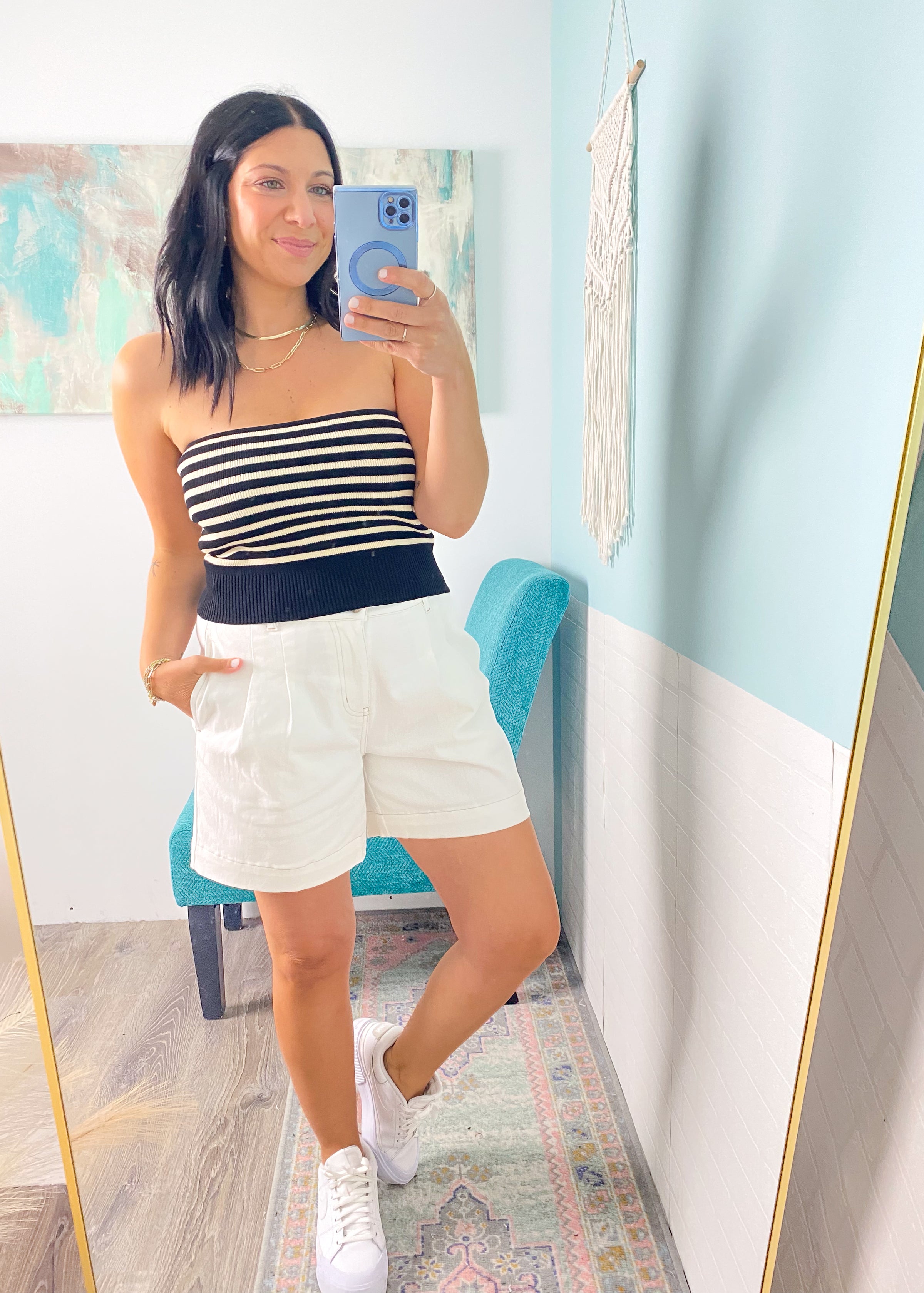 Woman taking a mirror selfie wearing a striped top and white shorts in a room with light blue walls.