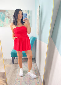 Woman taking a mirror selfie wearing a red strapless romper and white sneakers in a room with light blue walls.
