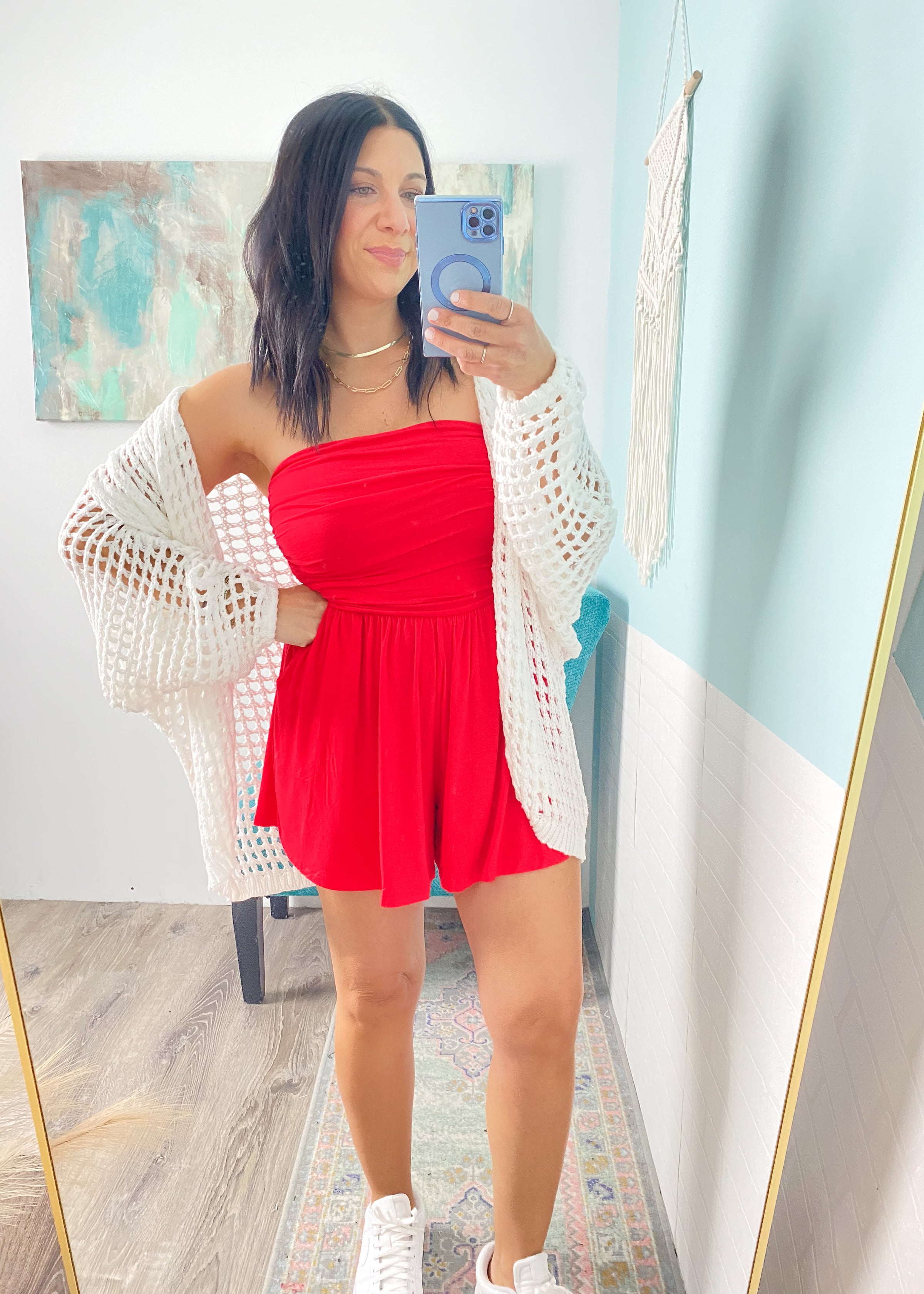 Woman taking a mirror selfie wearing a red dress and white  crochet cardigan in a room with light blue walls.