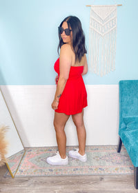 Woman in a red strapless romper  standing in a room with a white wall and decorative elements.