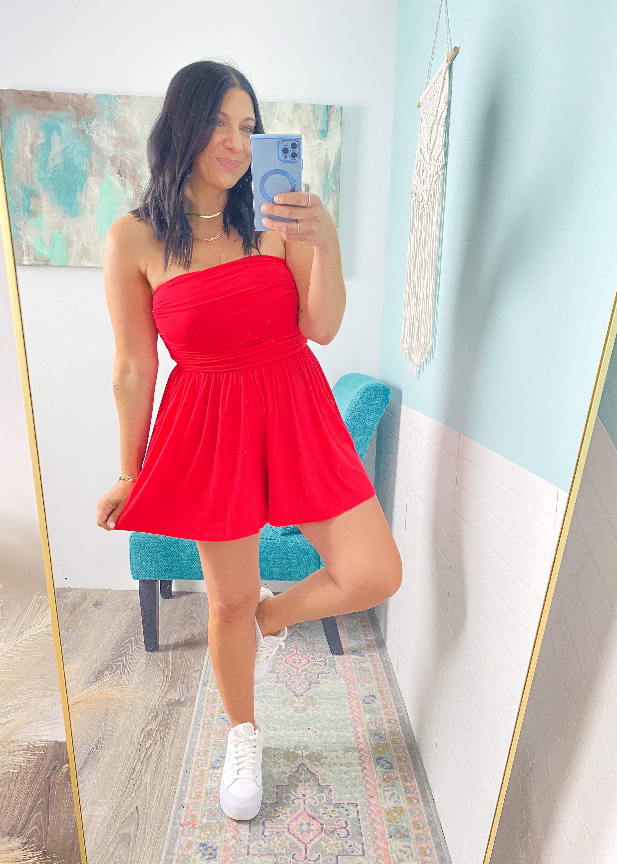 Woman taking a mirror selfie wearing a red strapless romper  in a room with light blue walls.