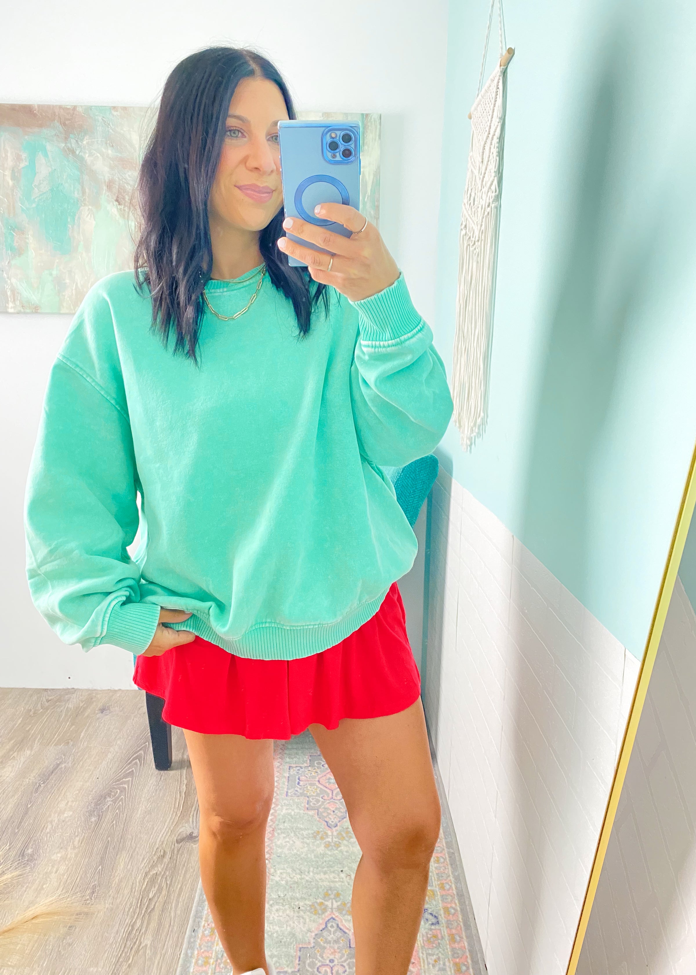 Woman taking a mirror selfie wearing a teal sweatshirt and red strapless romper  in a room with light blue walls.