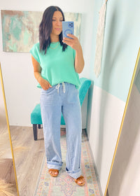 Woman taking a mirror selfie wearing a green top and light blue jeans in a room with a colorful abstract painting on the wall.