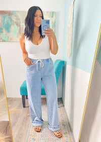 Woman taking a mirror selfie wearing a white tank top and light blue jeans in a room with a colorful rug and abstract painting.