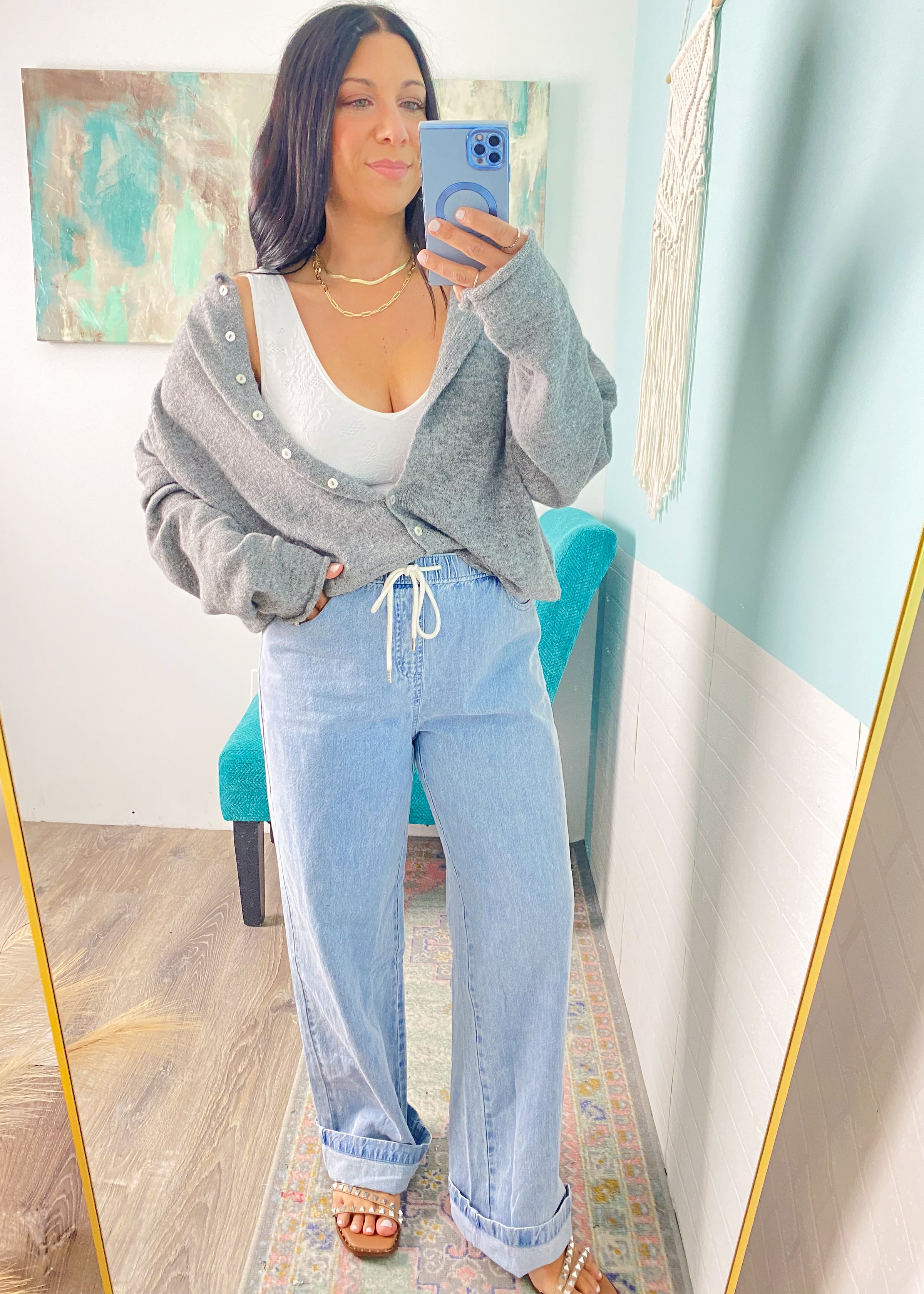 Woman taking a mirror selfie wearing a gray cardigan, white top, and light blue jeans.