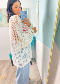 Woman wearing a white crochet cardigan top and light blue jeans, standing in a room with a colorful painting on the wall.