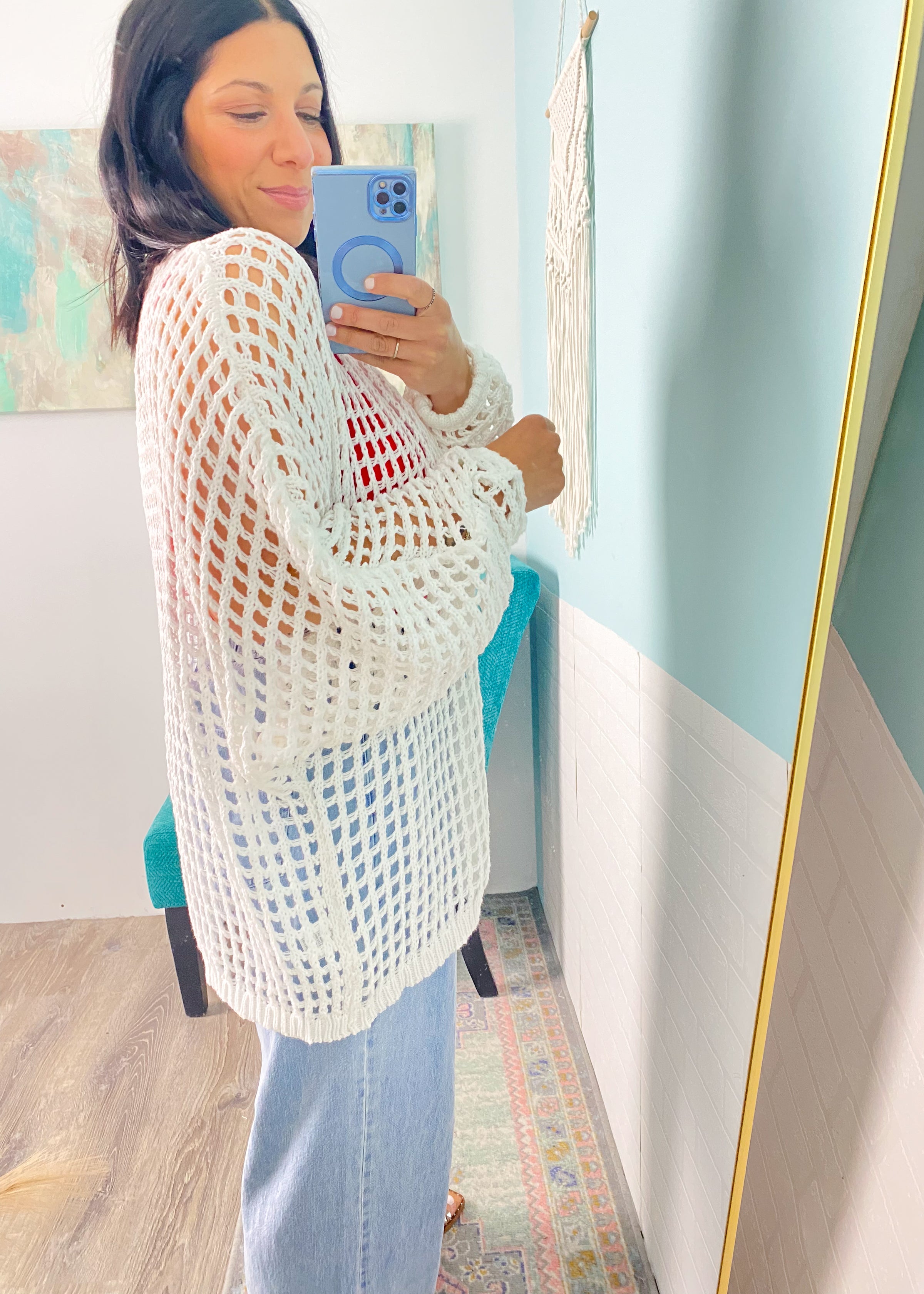 Woman wearing a white crochet cardigan top and light blue jeans, standing in a room with a colorful painting on the wall.