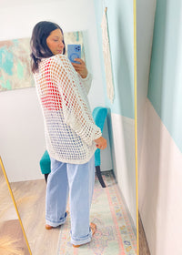 Woman taking a mirror selfie wearing a white crochet cardigan  and light blue jeans in a room with colorful decor.