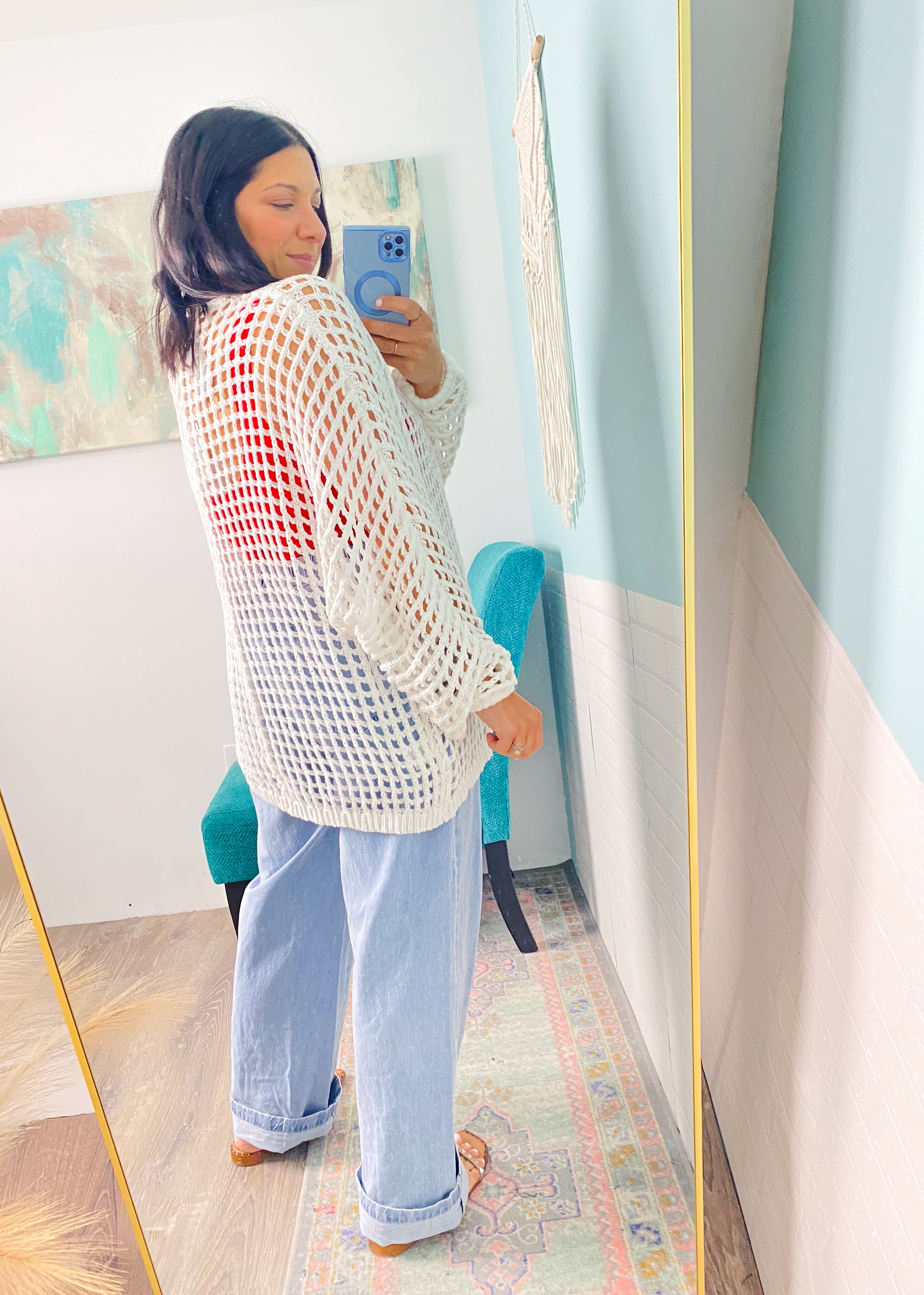 Woman taking a mirror selfie wearing a white crochet cardigan  and light blue jeans in a room with colorful decor.