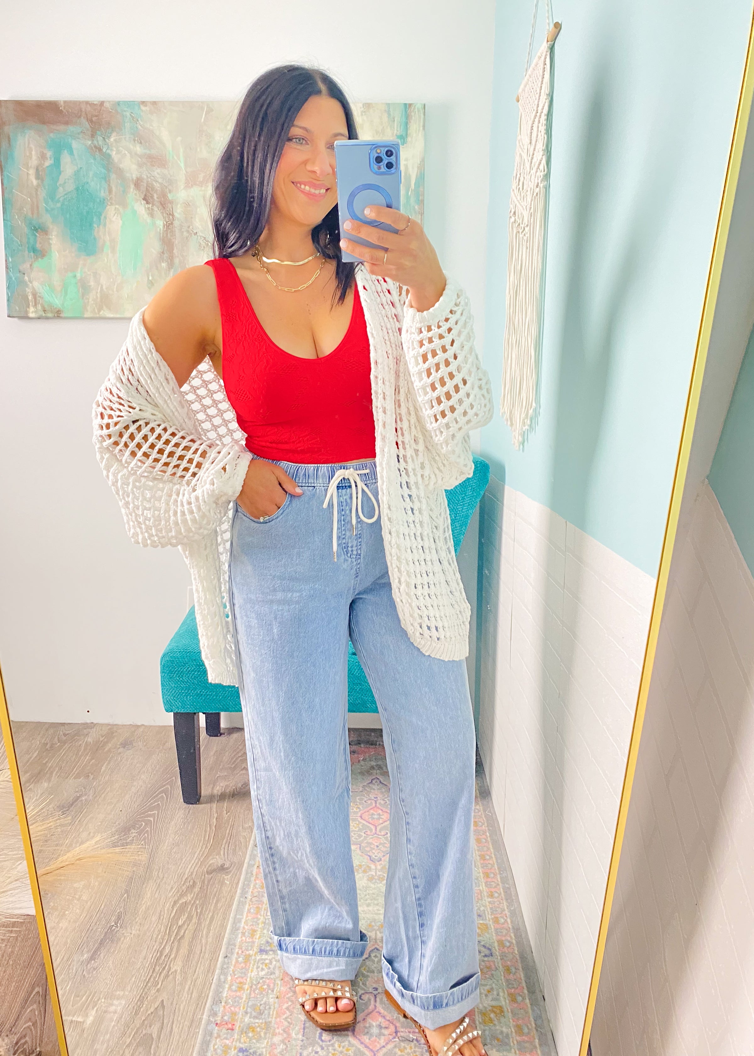 Woman taking a mirror selfie wearing a red top, light blue jeans, and a white crochet cardigan.