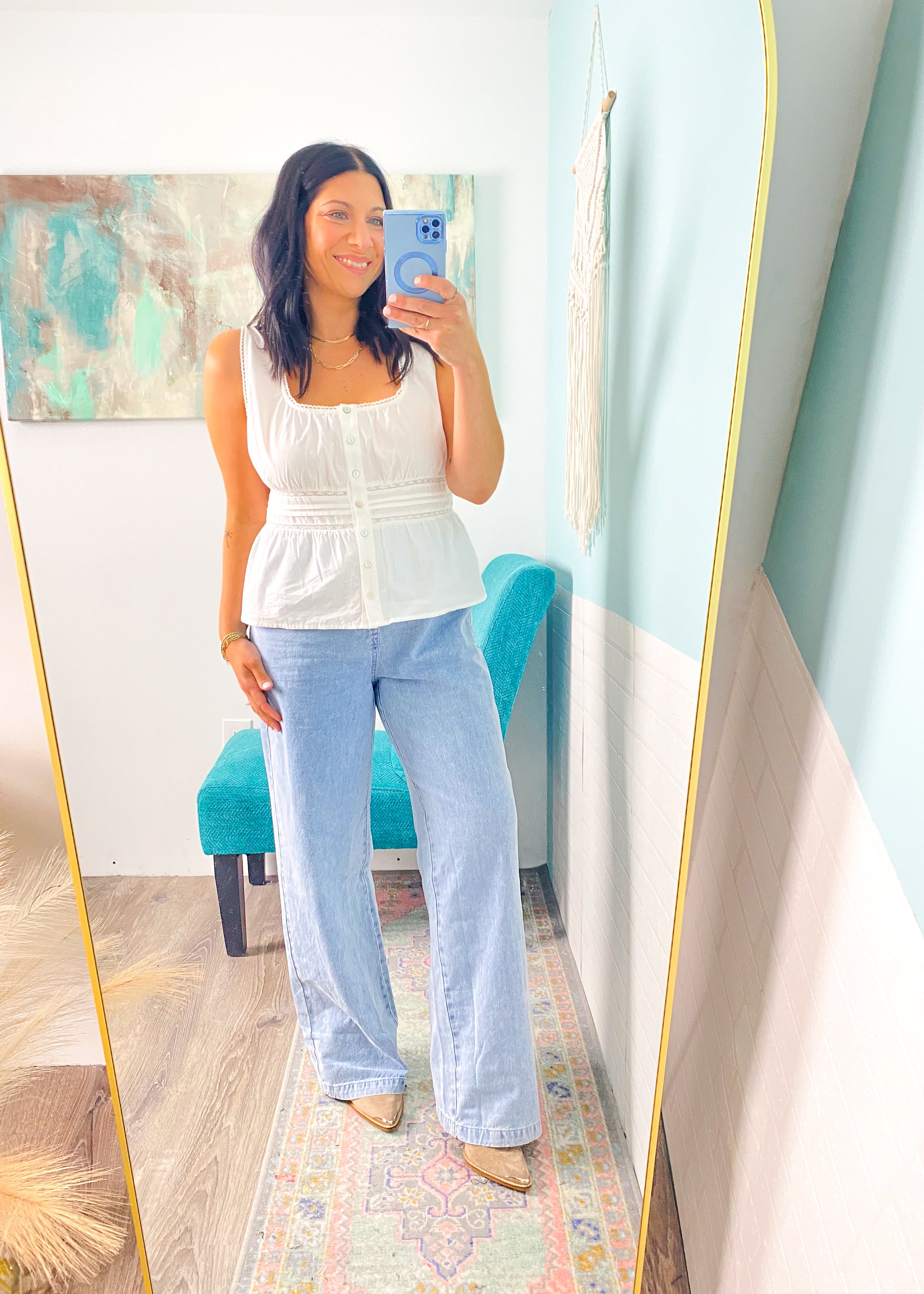 Woman taking a mirror selfie wearing a white peplum top and light blue jeans in a room with a colorful rug and teal chair.