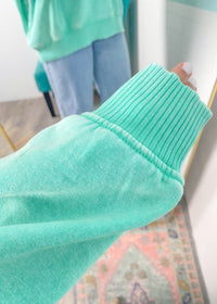 Close-up of a person wearing a mint green sweatshirt showing the ribbed wrist cuff with a blurred indoor background