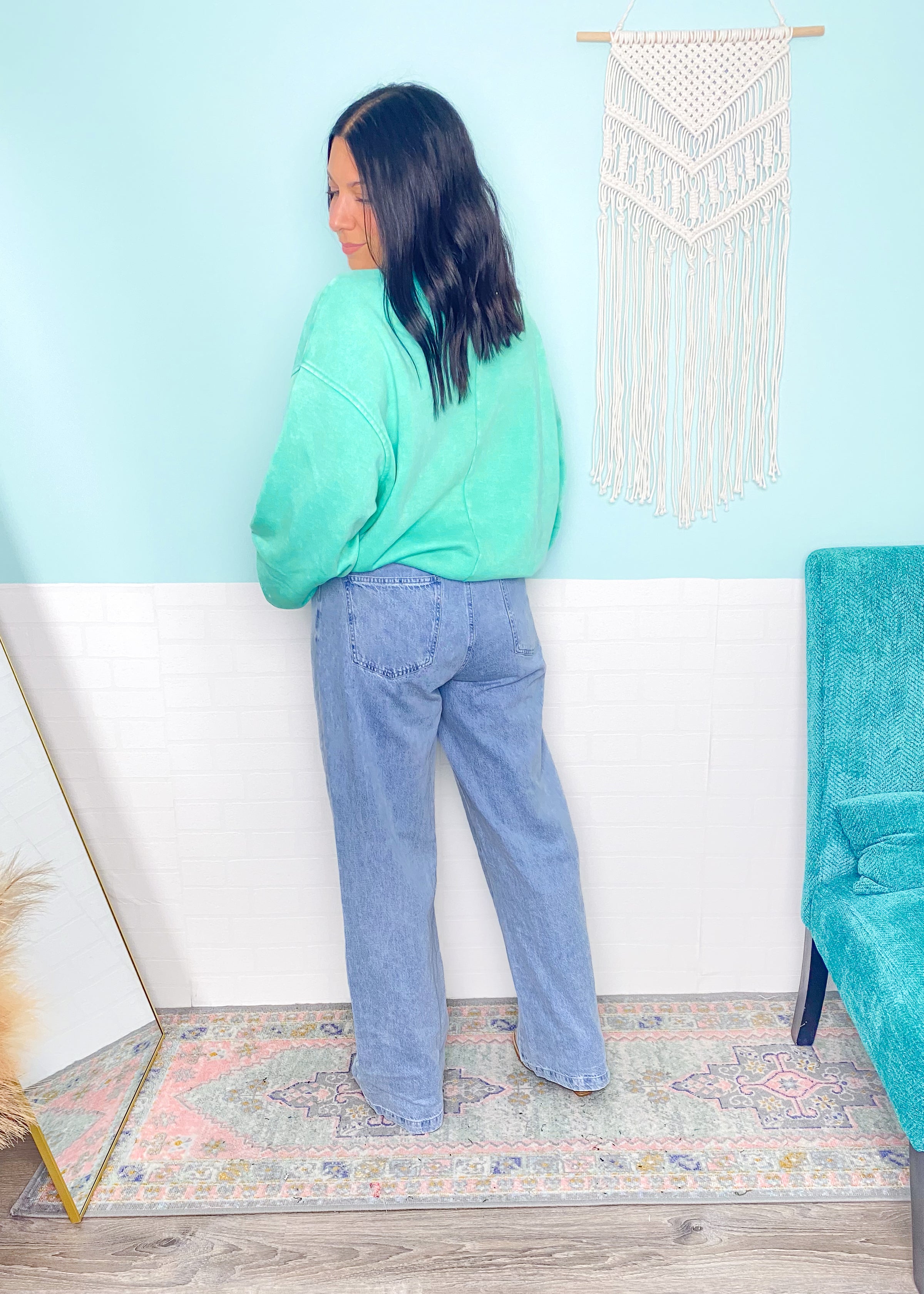 Person wearing a green sweater and blue jeans standing in a room with a light blue wall and decorative elements.