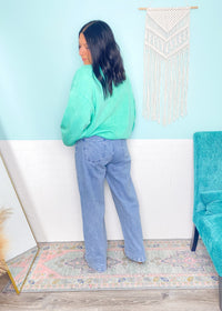 Person wearing a mint green sweathirt and blue jeans standing in a room with a light blue wall and decorative elements.