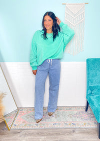 Woman wearing a teal sweatshirt and blue jeans standing in a room with a white wall and decorative elements.
