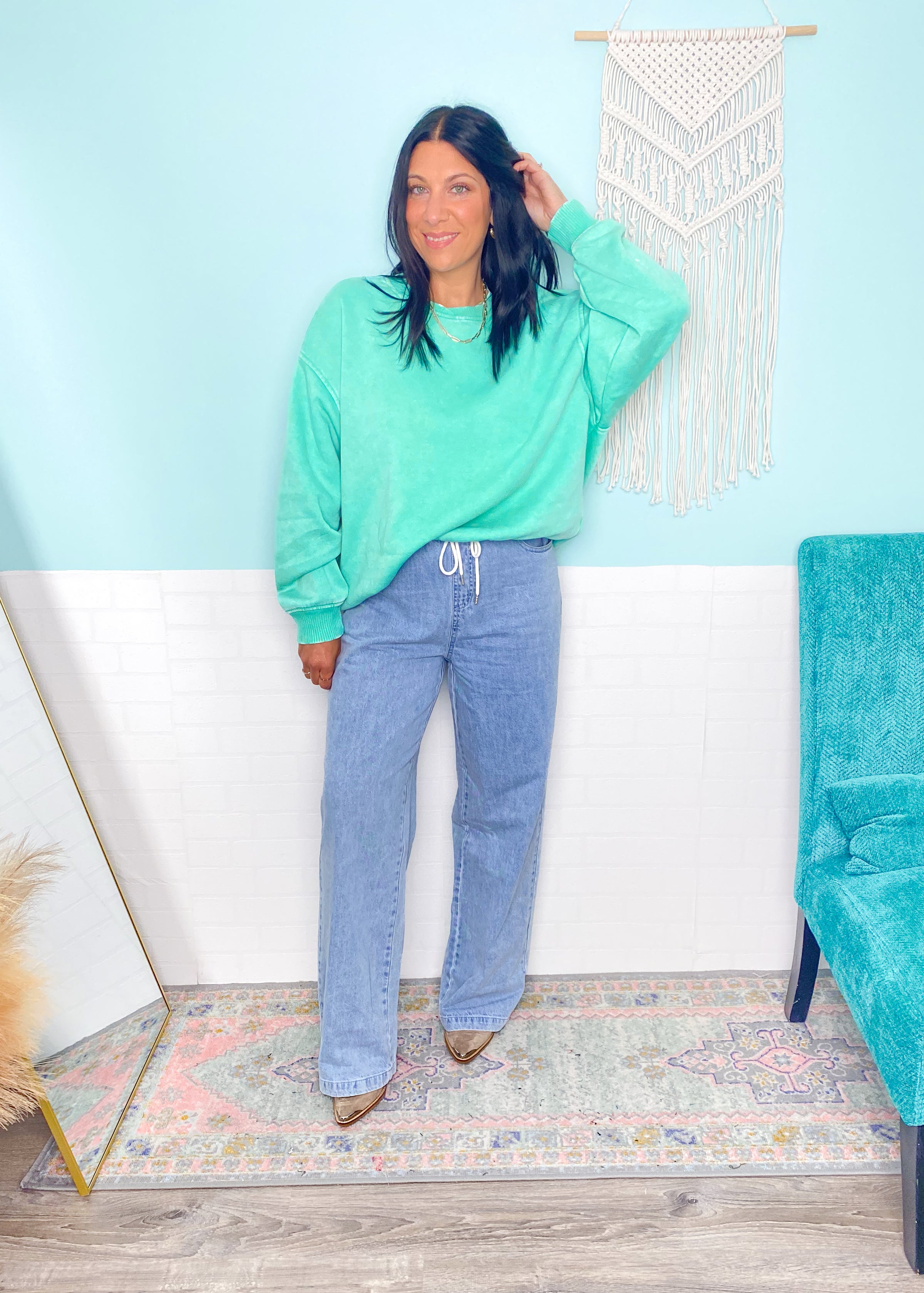 Woman wearing a teal sweatshirt and blue jeans standing in a room with a white wall and decorative elements.