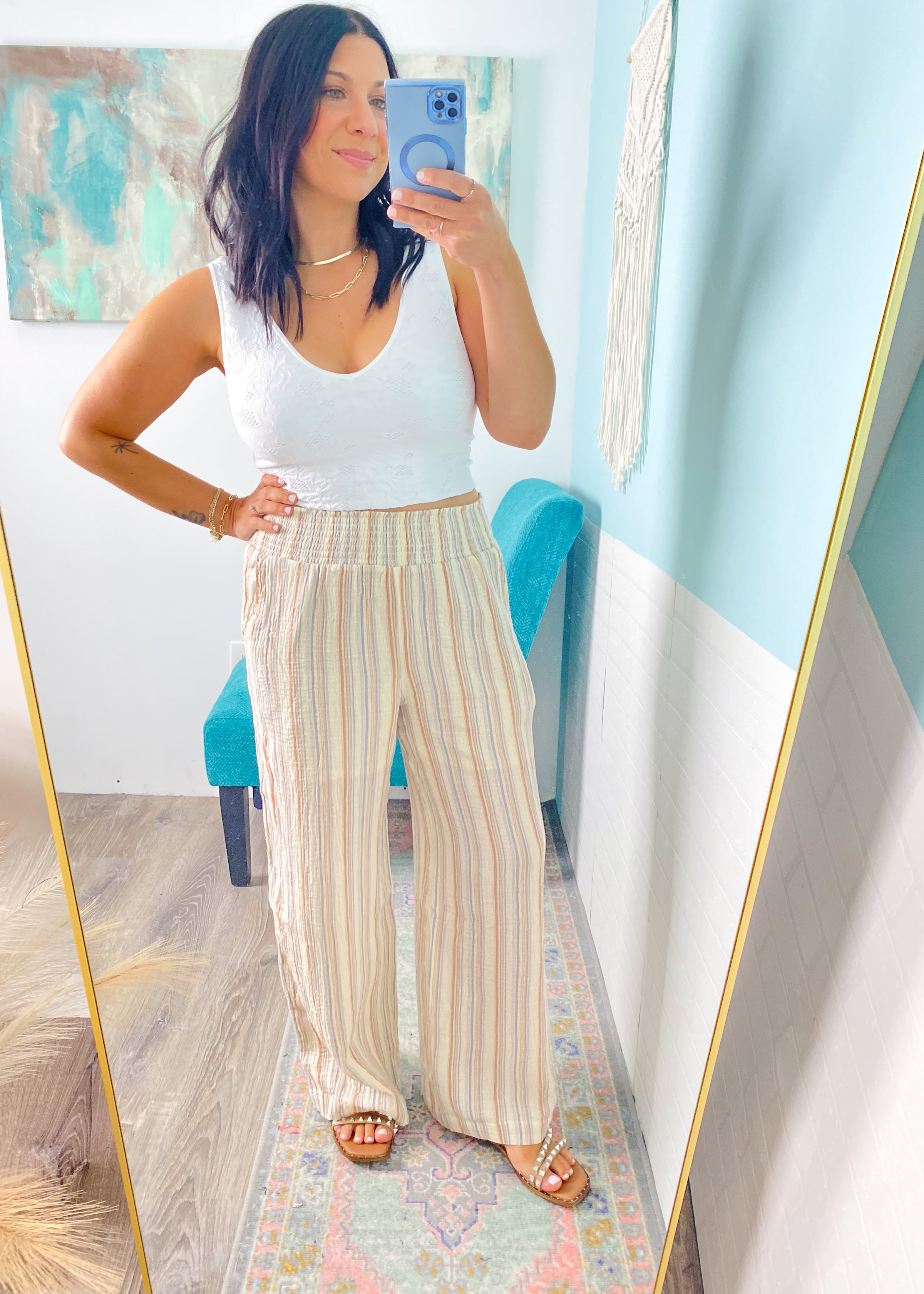 Woman taking a mirror selfie wearing a white lace patterned seamless cropped top and striped pants in a room with a colorful rug and blue chair.