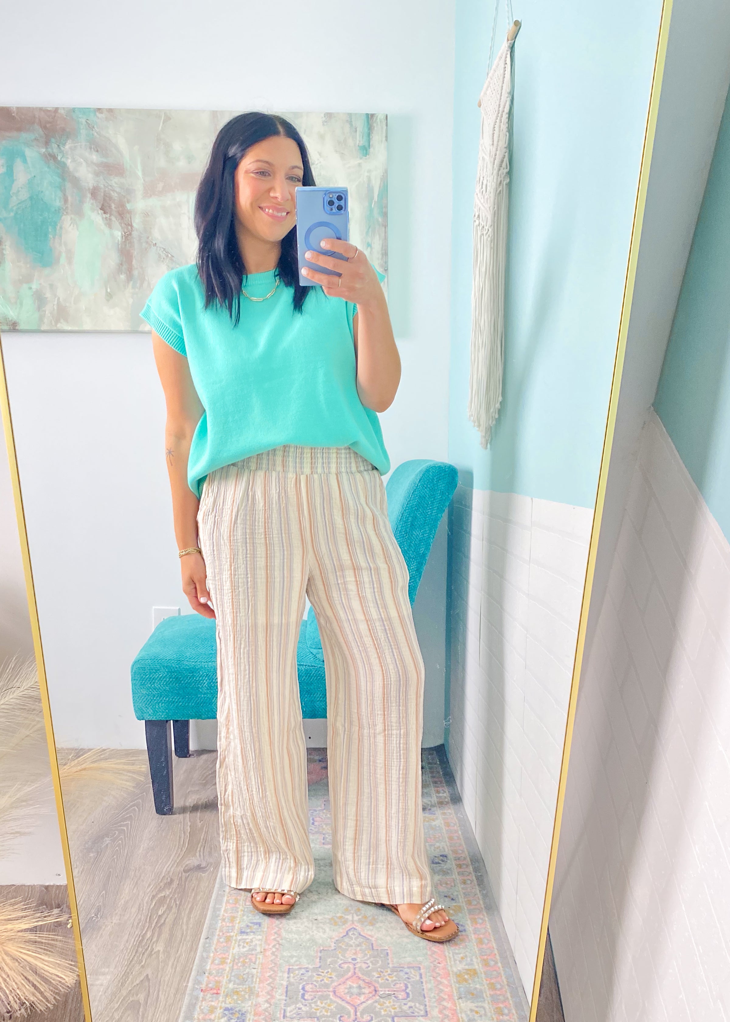 Woman taking a mirror selfie wearing a teal top and striped pants in a room with a blue wall.
