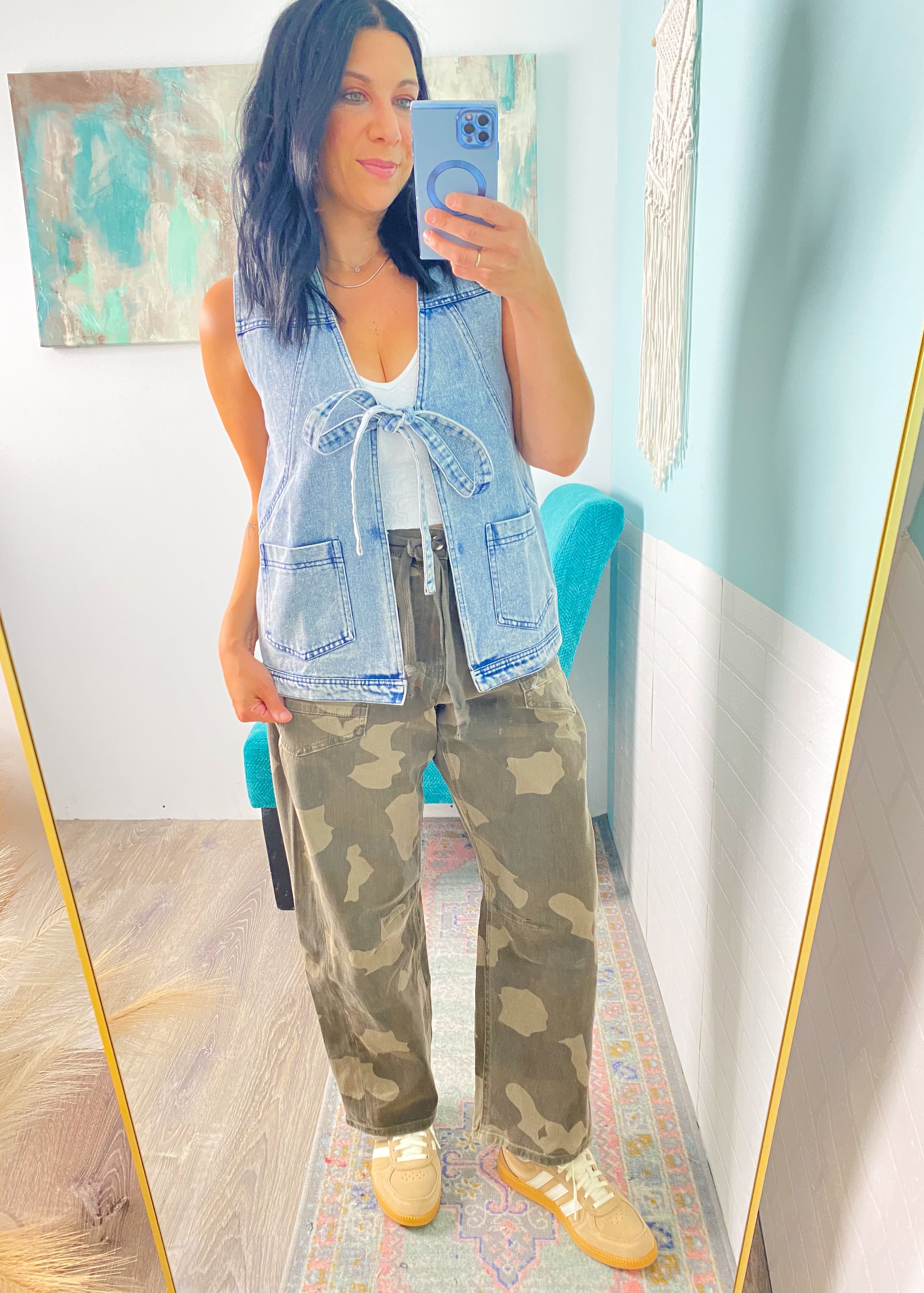 Woman taking a mirror selfie wearing a denim vest, white top, camouflage pants, and yellow shoes.