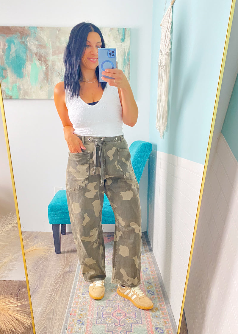 Person taking a mirror selfie wearing a white tank top and camouflage pants in a room with a colorful rug and blue wall.