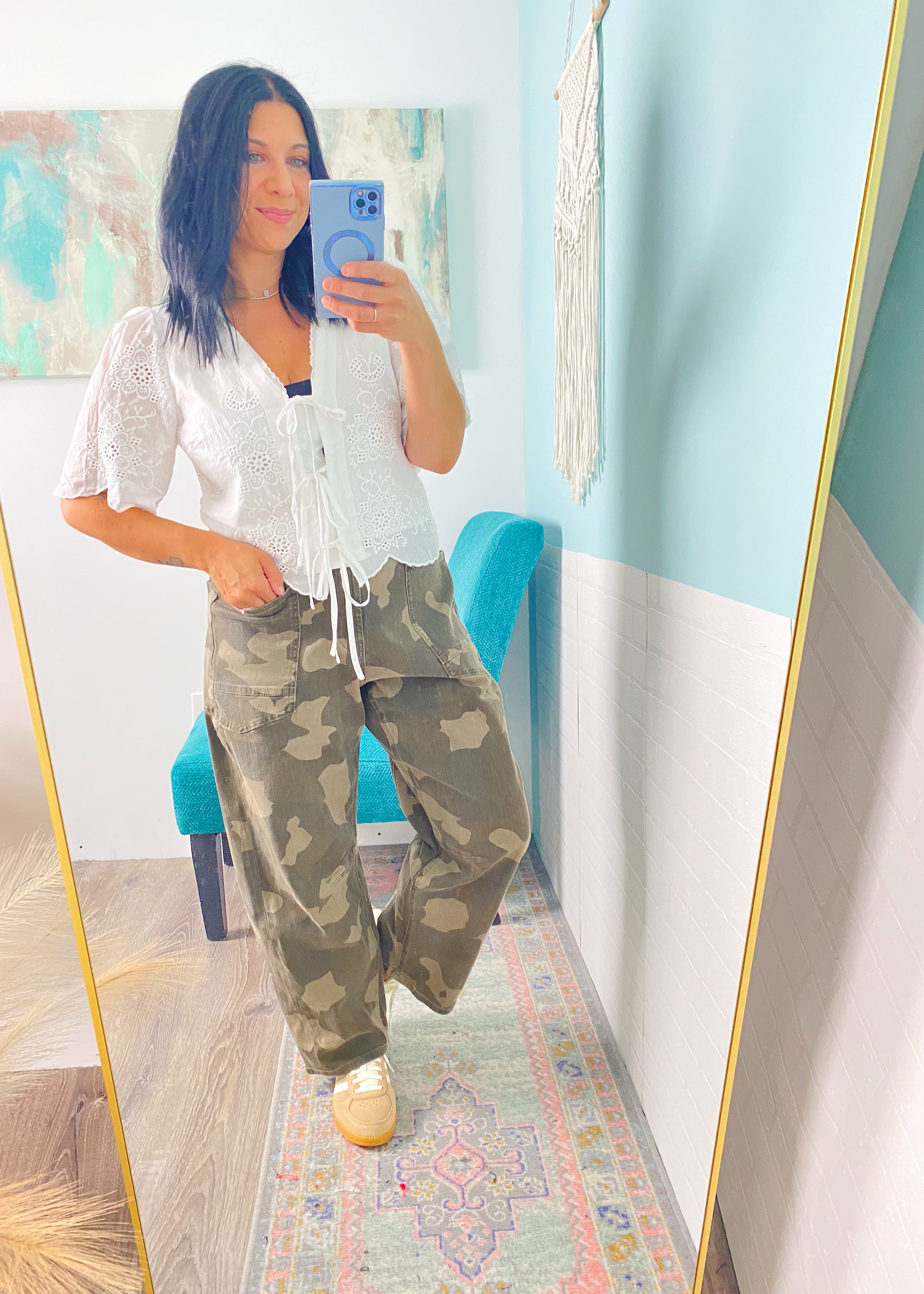 Woman taking a mirror selfie wearing a white top and green camouflage pants in a room with light blue walls.